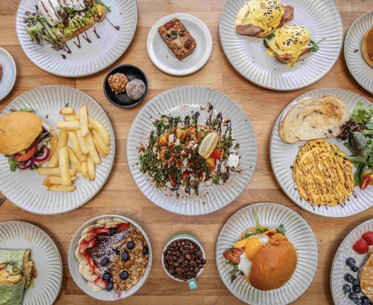 Overhead spread of Awaken Avalon dishes — avocado toast, eggs benedict, burger and chips, acai bowl, omelette and coffee across a timber table